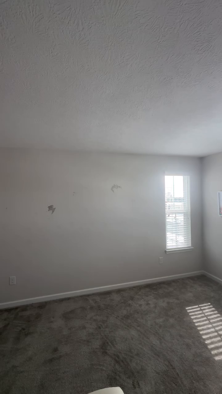 Empty room with gray carpet, white walls with damage, and a window.