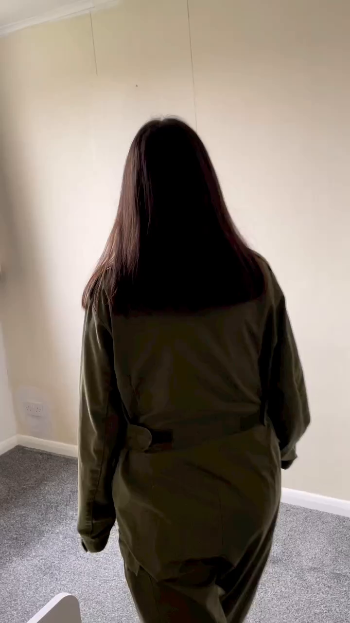 A person with long brown hair wearing a belted green jumpsuit.