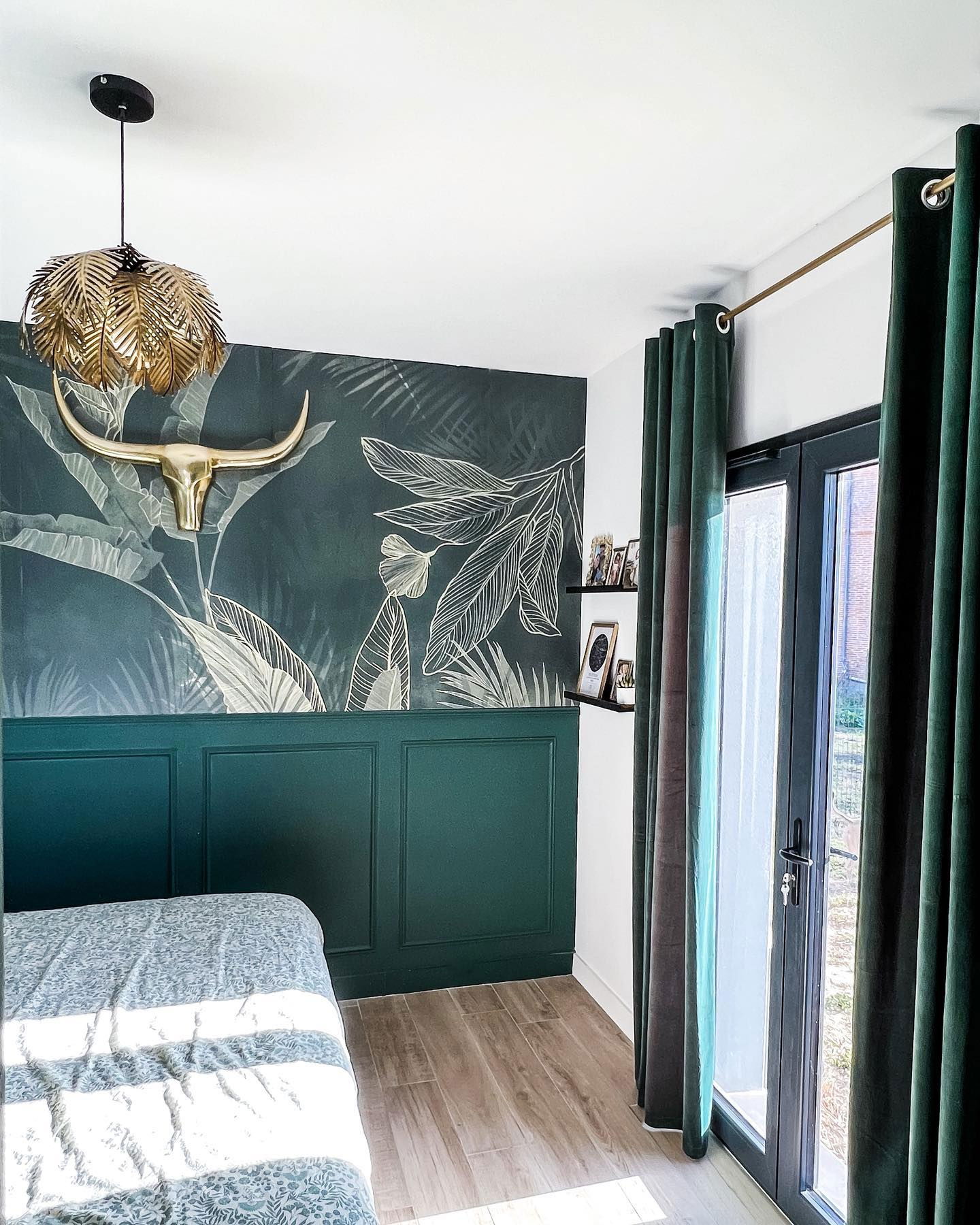 Bedroom with tropical wallpaper, gold bull skull, and leaf-shaped pendant light. Green wainscoting and curtains.