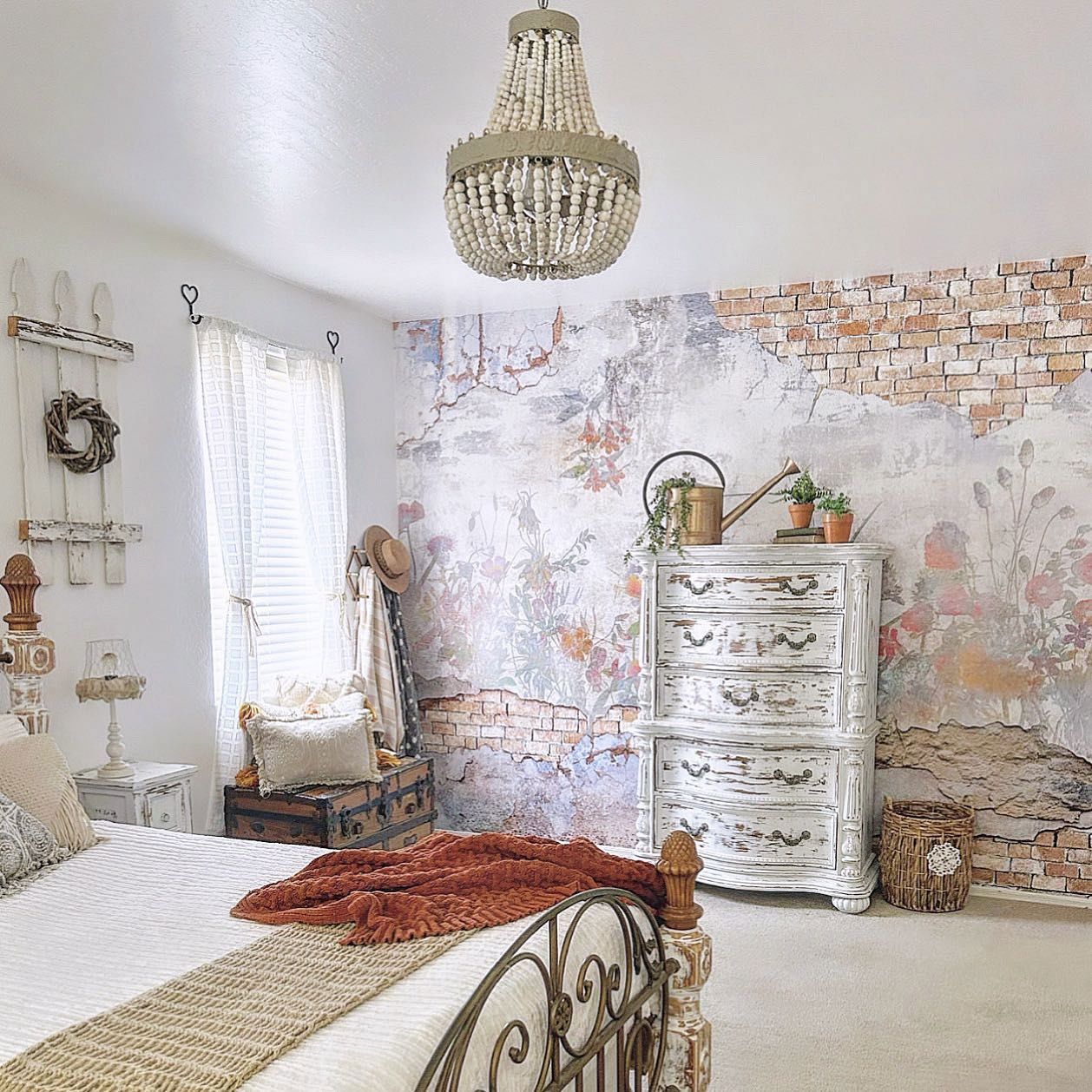 Bedroom with floral brick wallpaper, beaded chandelier, and distressed white dresser.