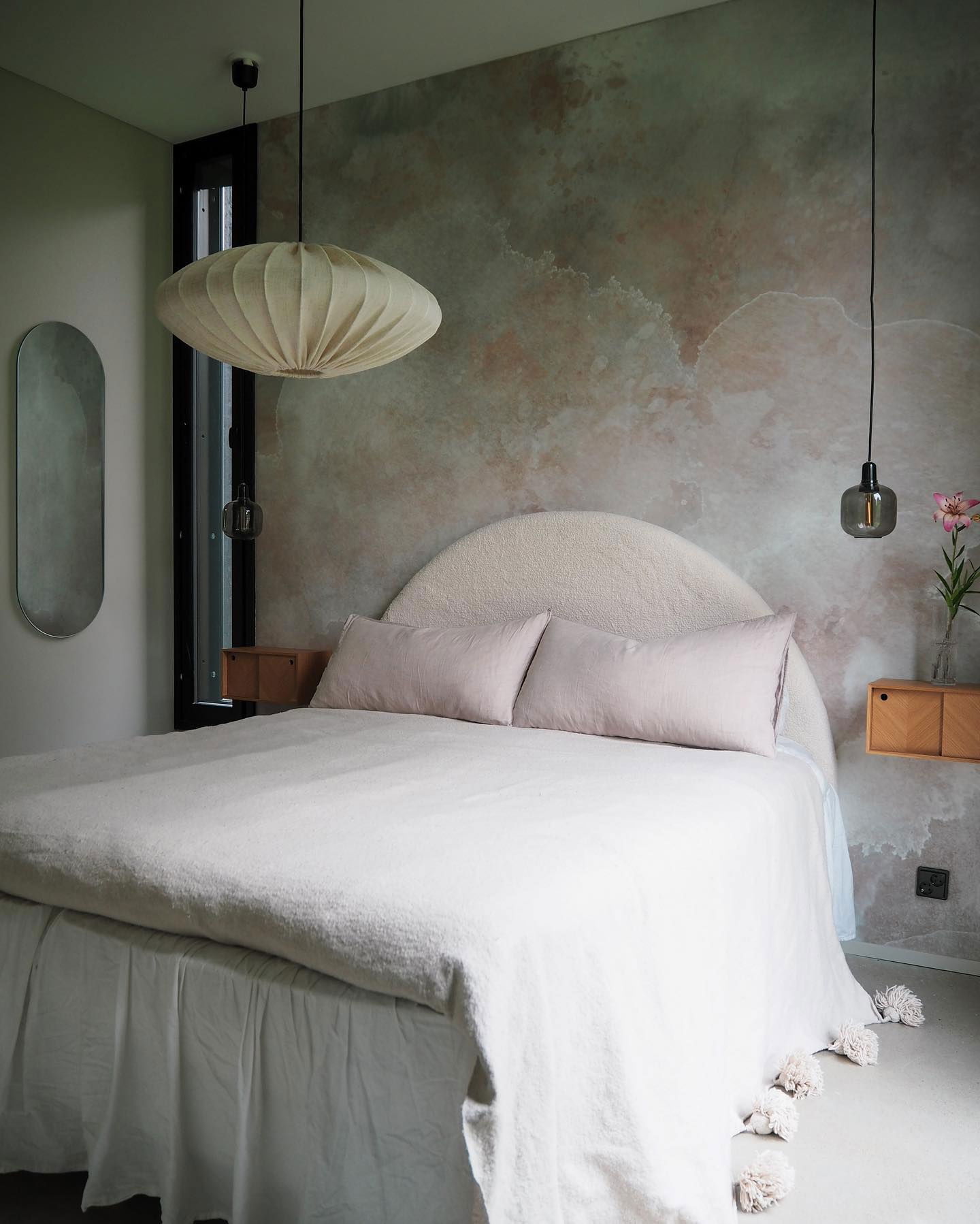 Dormitorio con pared texturizada, cama con ropa de cama blanca y ribete de pompón, y lámparas colgantes.