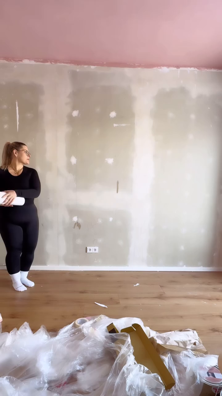 A woman in a black jumpsuit holds a roll of wallpaper in a room being renovated.