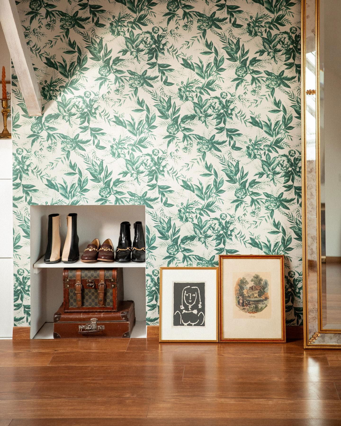 Interno con carta da parati floreale, pavimento in legno e scaffale a muro. Sono esposti stivali, valigie e opere d'arte incorniciate.