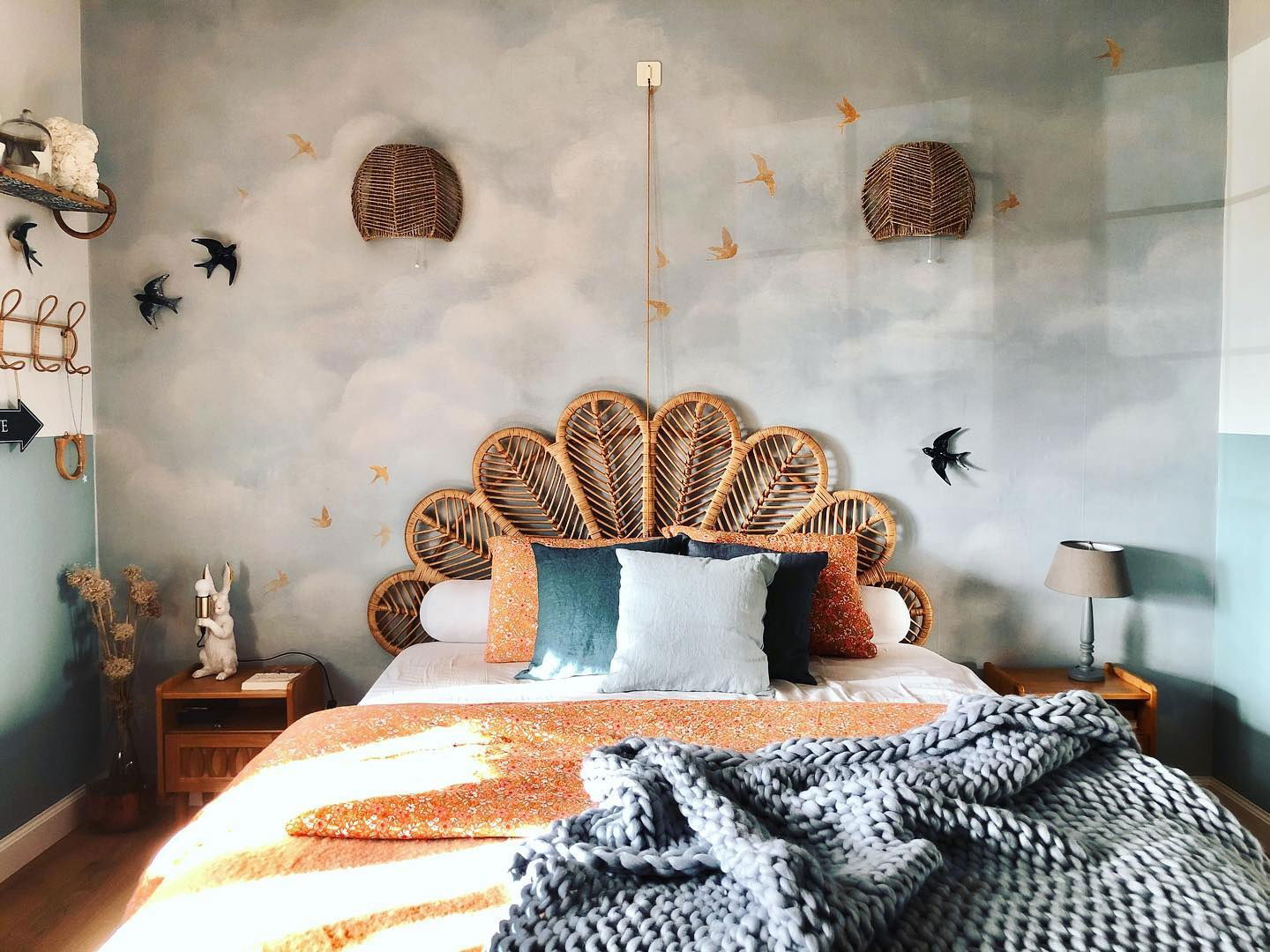 Bedroom with rattan headboard, cloud wallpaper, and knit blanket. Wicker sconces and bird wall decor add to the whimsical style.