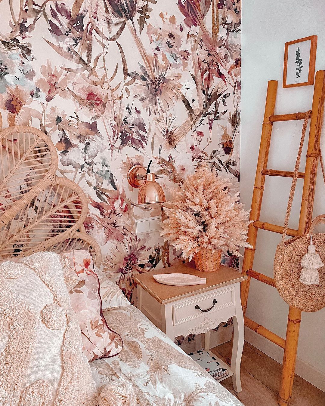 Mysigt sovrum med blommiga tapeter, sänggavel i rotting och bambustegen. Sängbord med torkade blommor och kopparlampa. Vävd väska hänger på stegen.