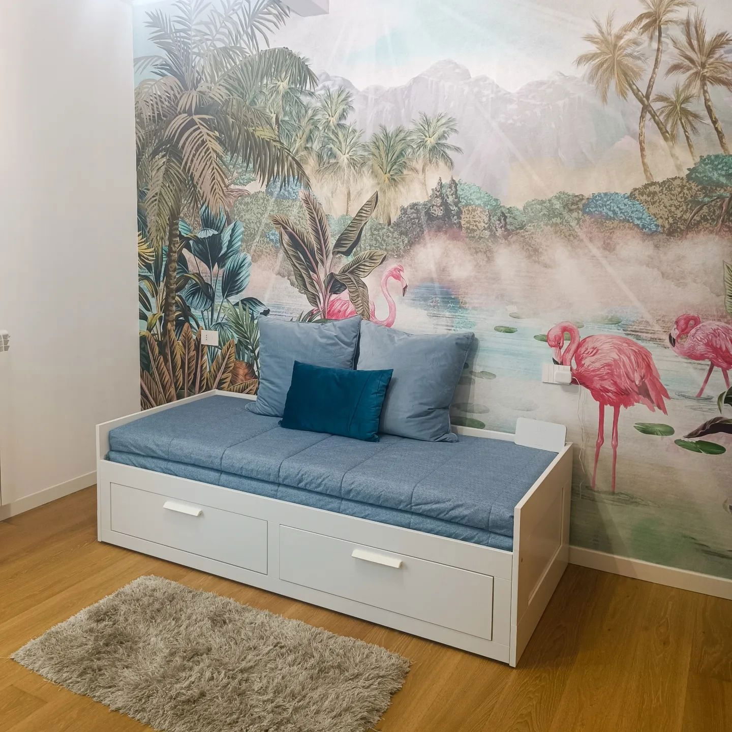 Bedroom with white daybed, blue cushions, and tropical flamingo wallpaper.