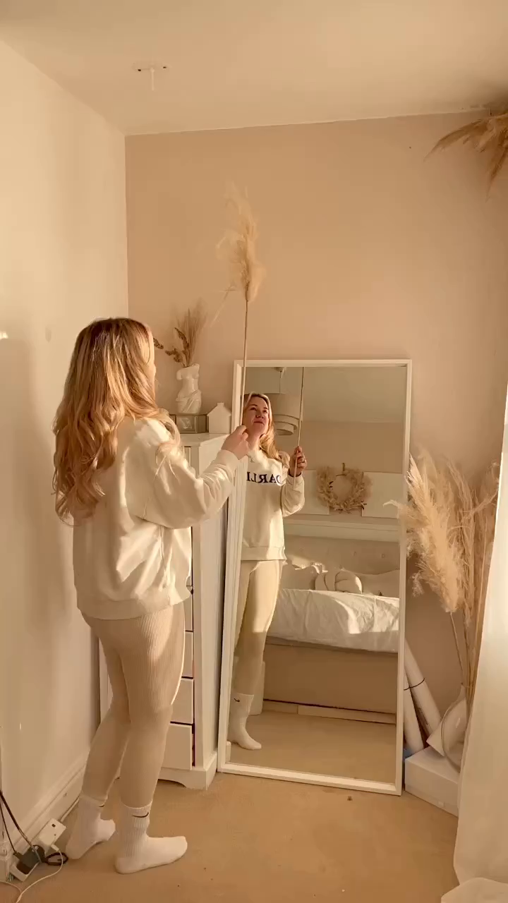 Woman in cream outfit holds pampas grass in front of mirror in bedroom.