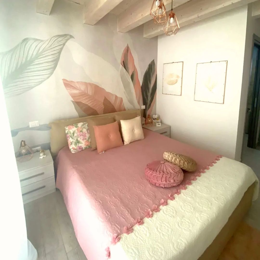 Camera da letto con biancheria da letto rosa, cuscini floreali e arte murale a tema foglia.