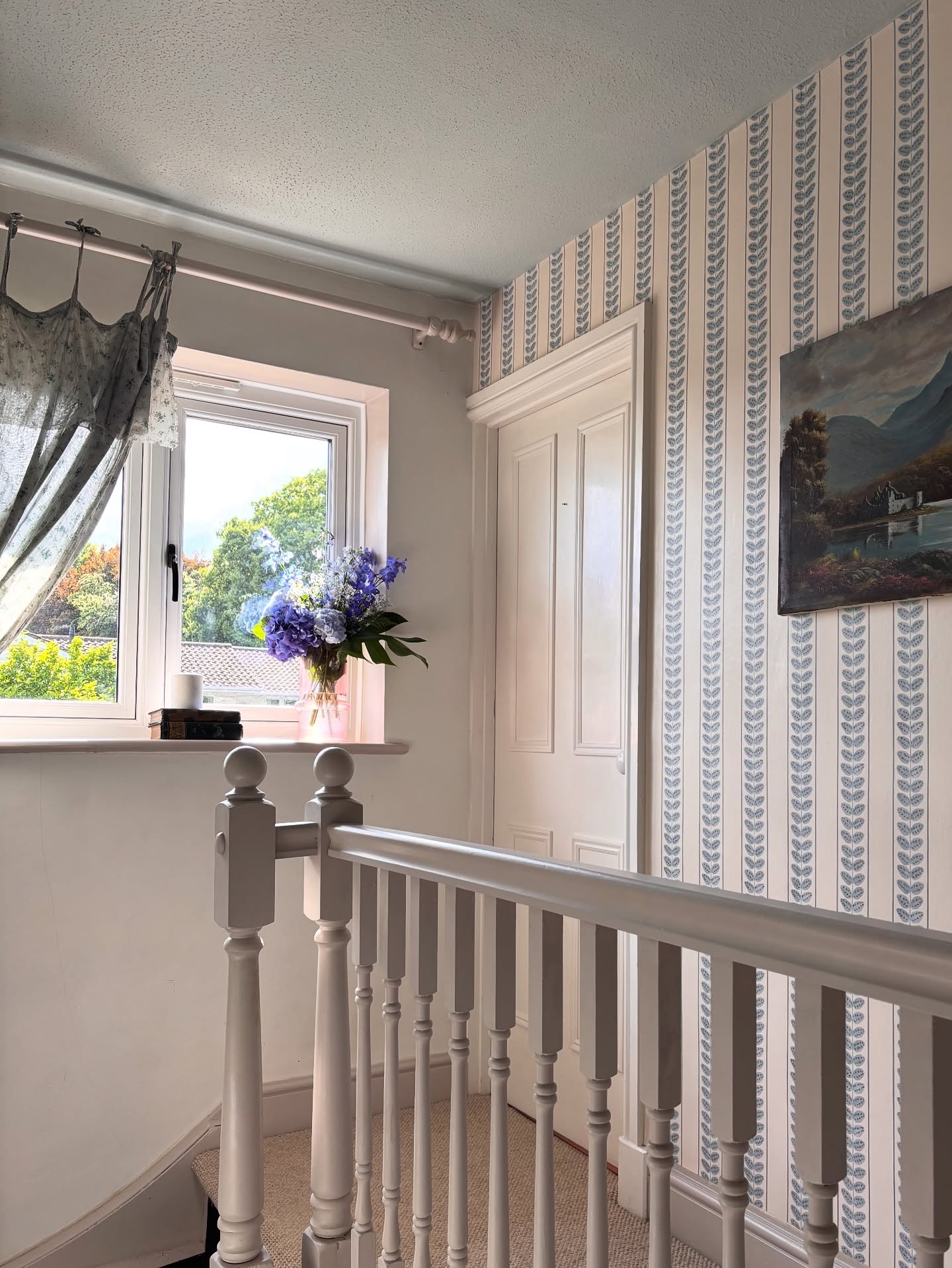 Interior view of a hallway with blue floral wallpaper and a gray painted banister. A window with flowers sits at the top of the stairs.