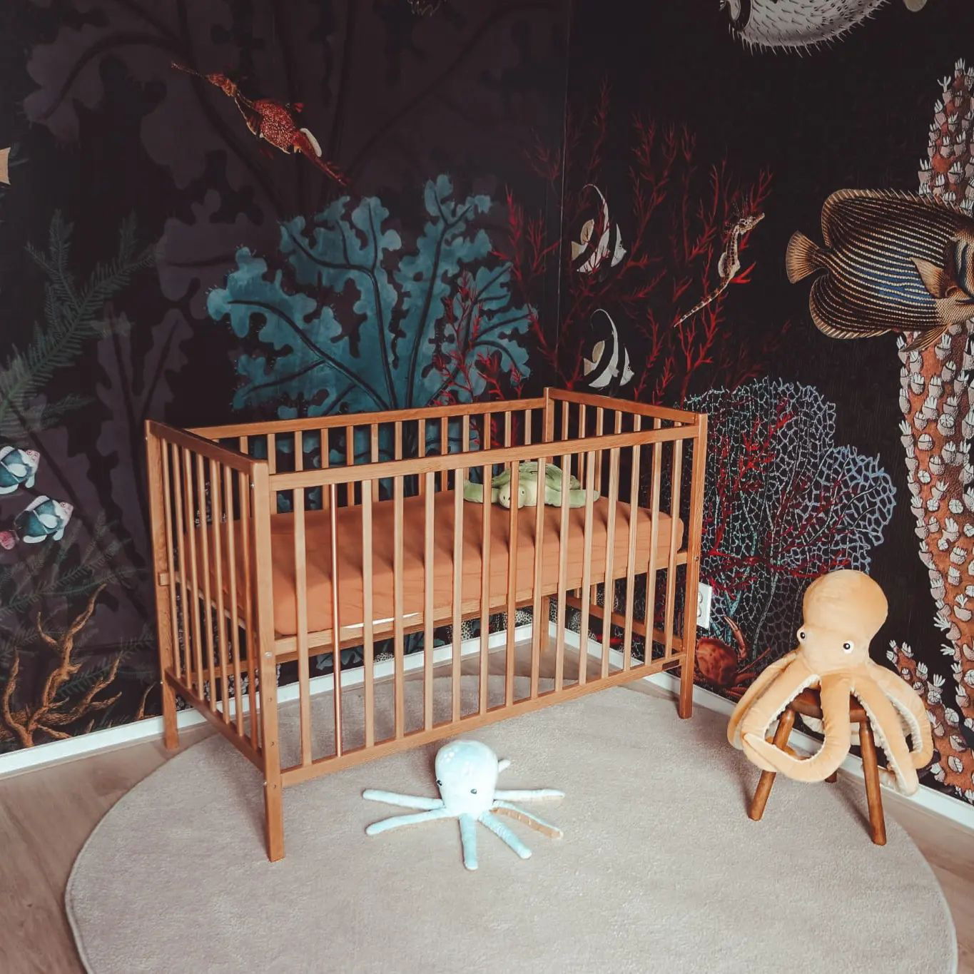 Nursery with wooden crib, sea-themed wallpaper, and octopus plush toys on a round rug.