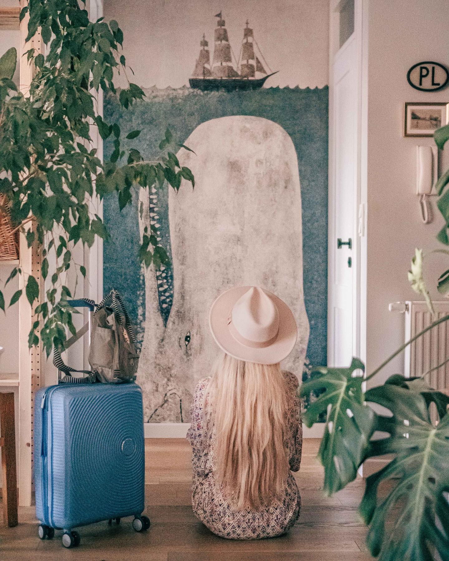Blonde woman in hat sits by blue suitcase in front of whale mural.