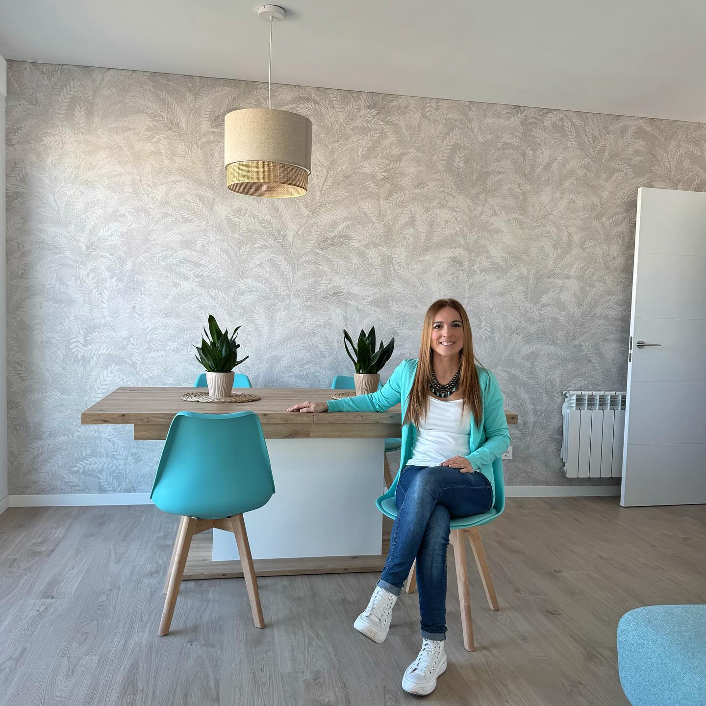 A woman sits at a dining table with turquoise chairs and potted plants.
