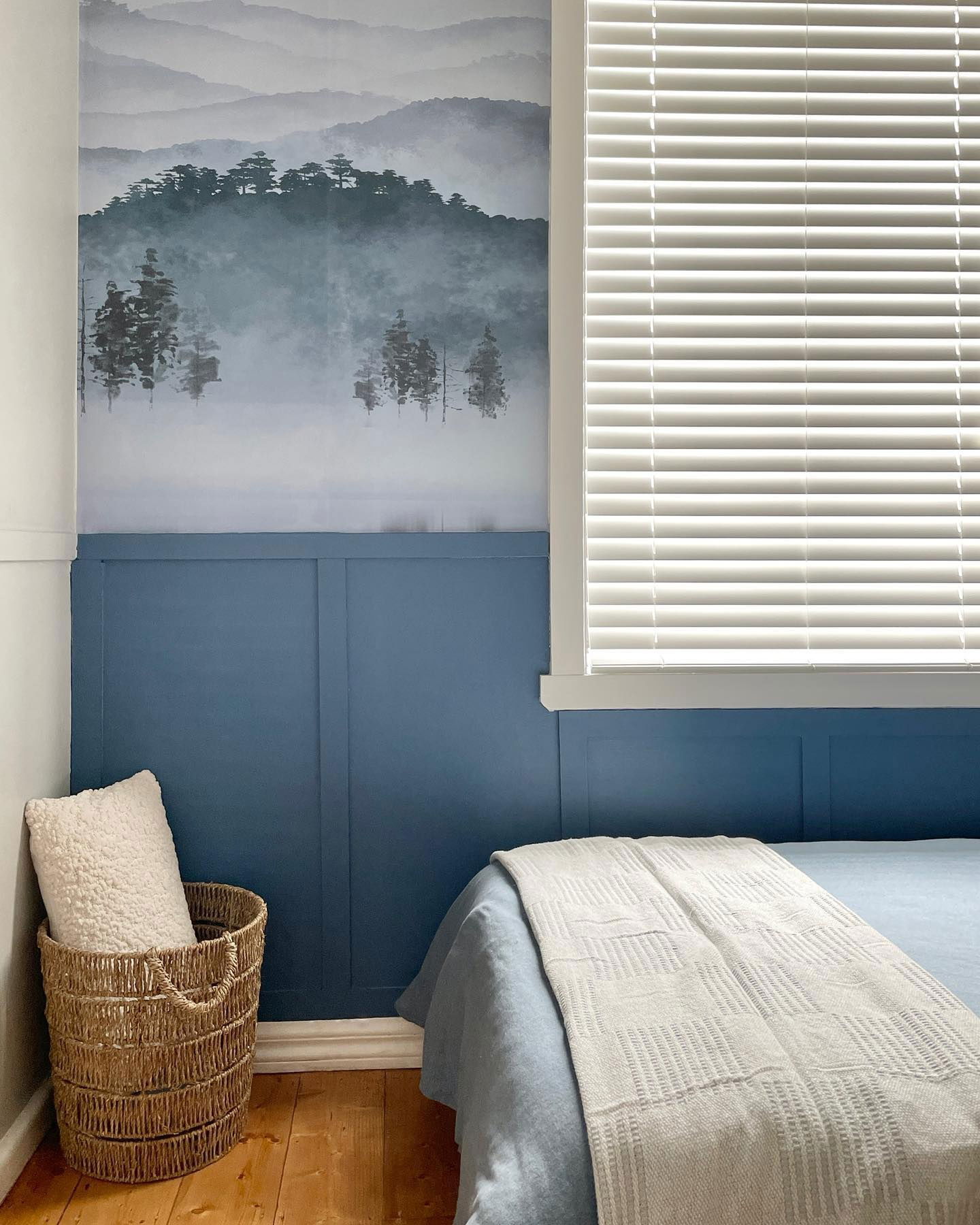 Schlafzimmerecke mit blau getäfelter Wand, Landschaftsgemälde und gewebtem Korb mit weißem Kissen.
