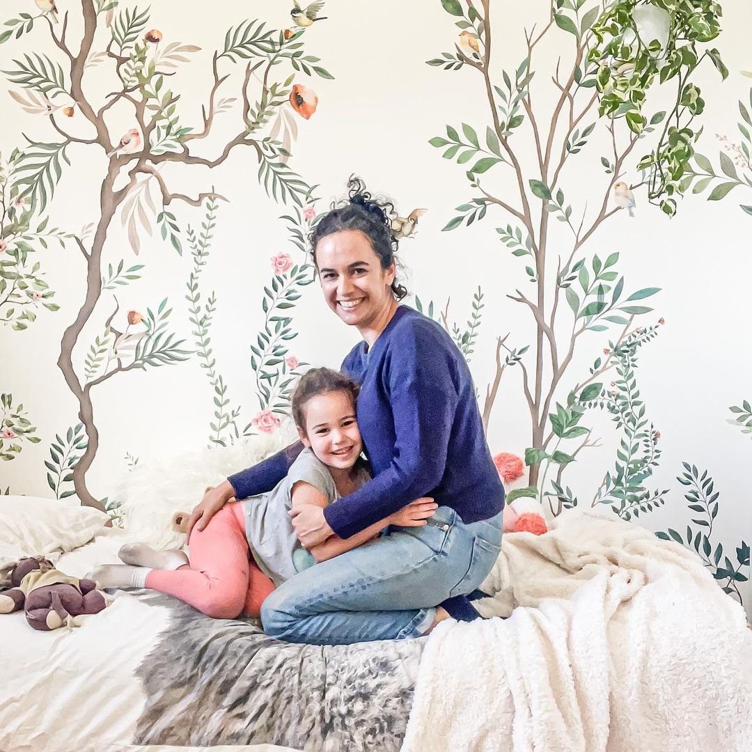 A smiling mother and daughter embrace on a bed with a floral mural.