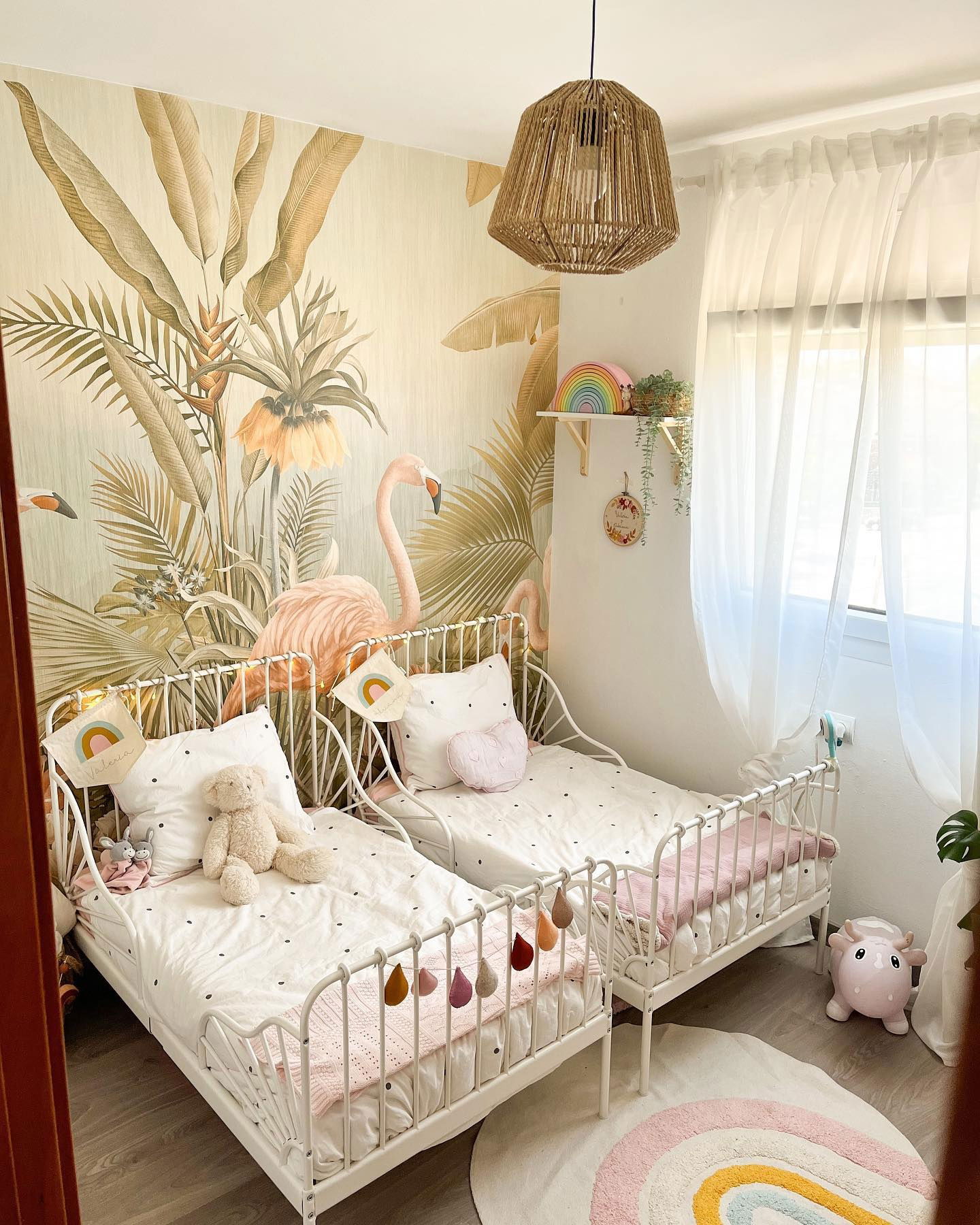 Dormitorio infantil con dos camas de metal blanco, papel pintado de flamencos, alfombra de arcoíris y lámpara colgante tejida.