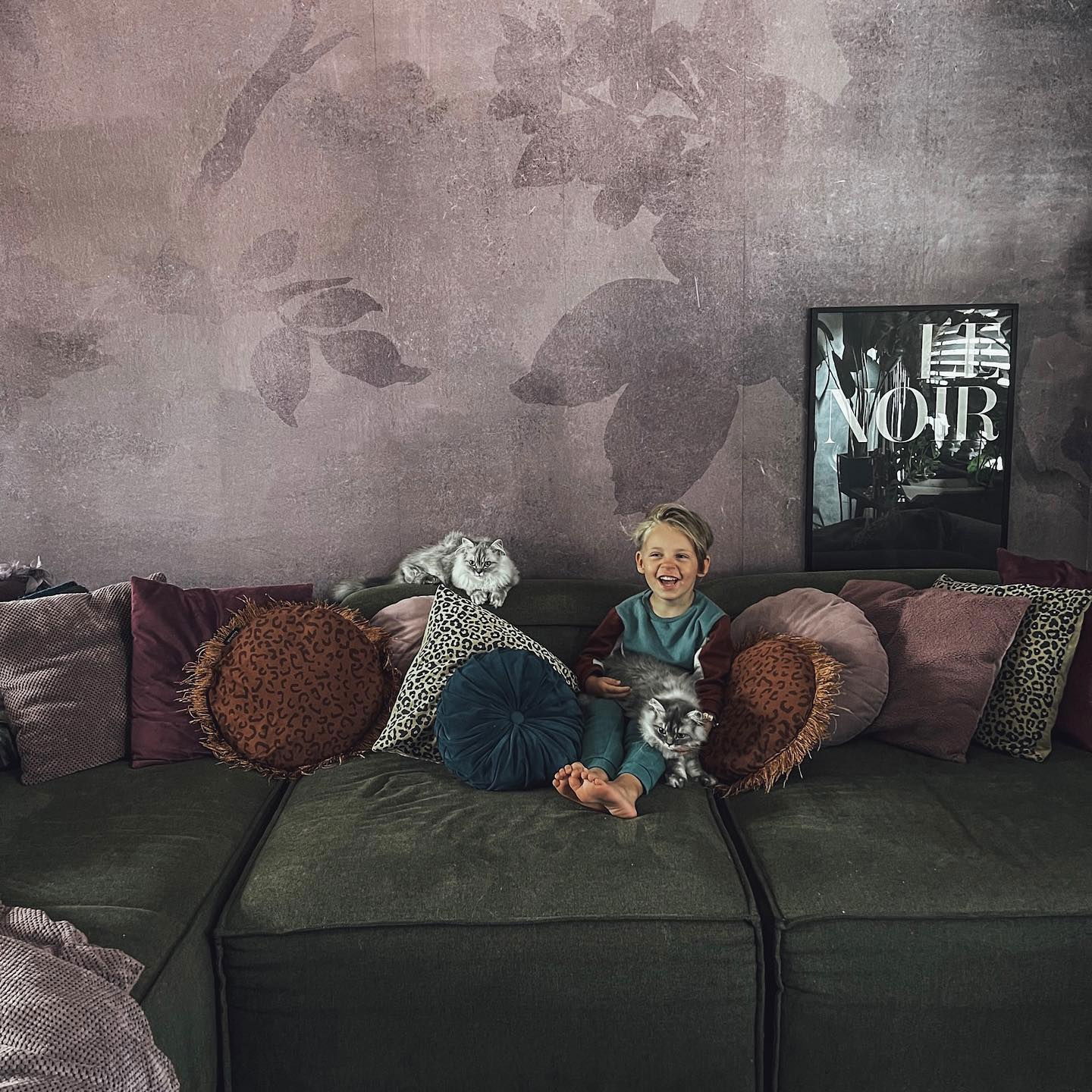 Et smilende barn sitter på en grønn sofa med to katter og mange puter.