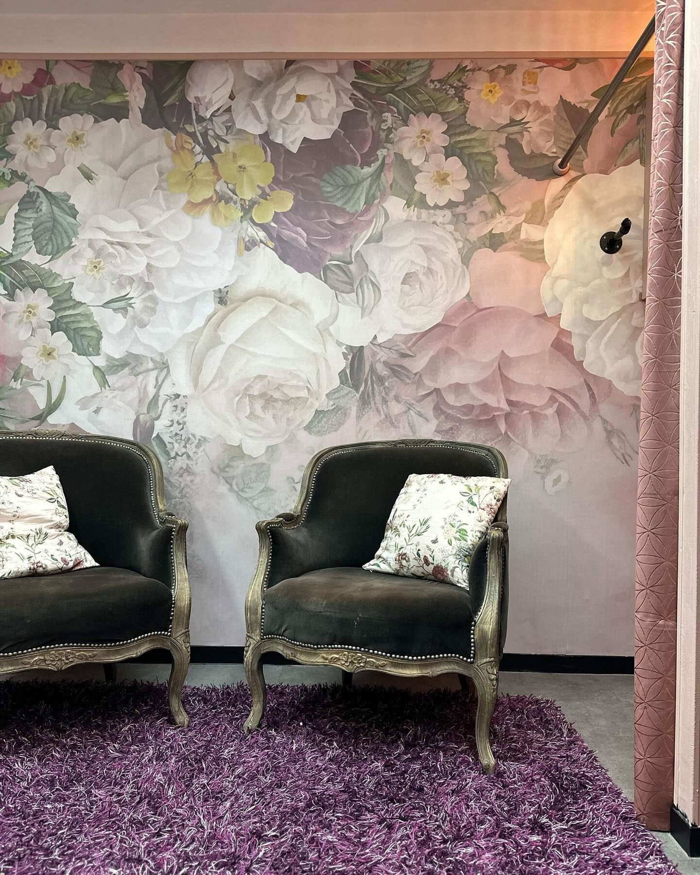 Interior with floral wallpaper, two dark armchairs with pillows, and a purple shag rug.