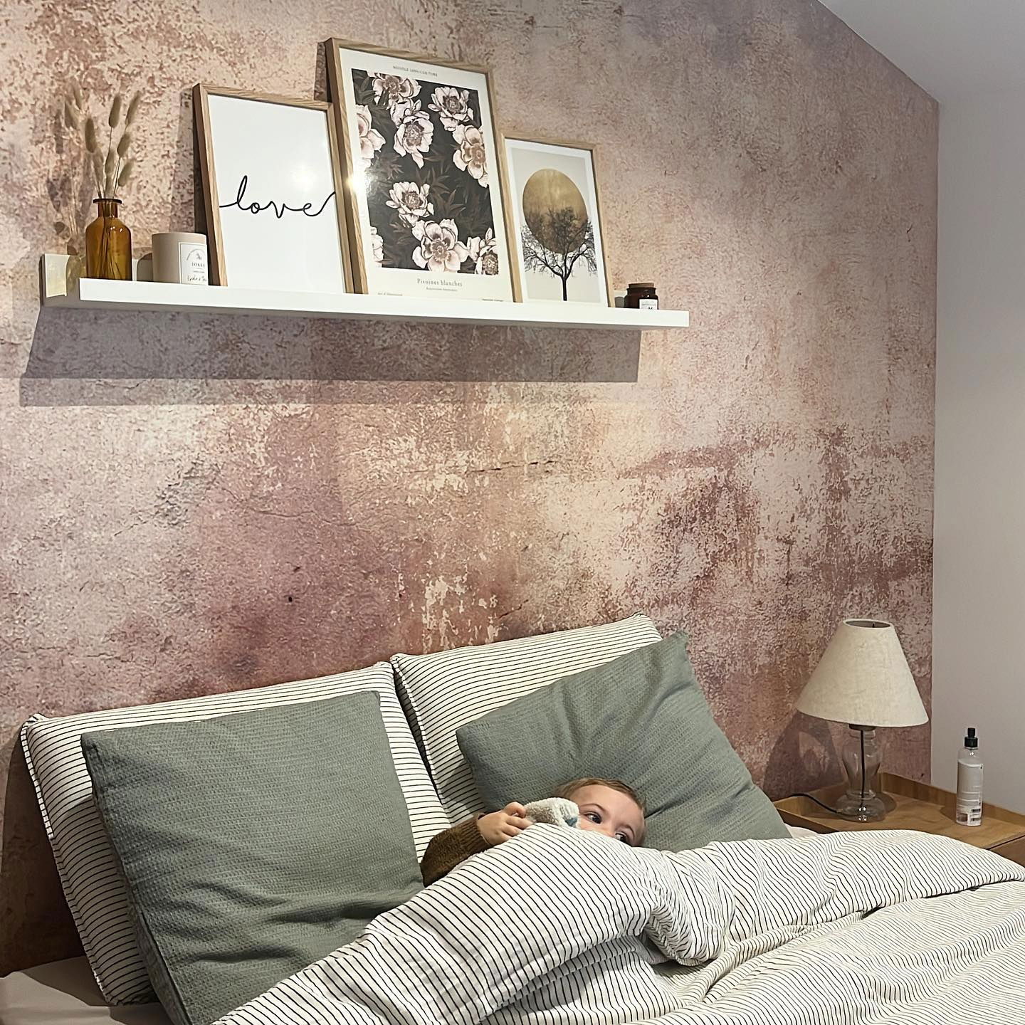 Bedroom scene with child peeking from striped bedding, textured wall, and framed art on a shelf.