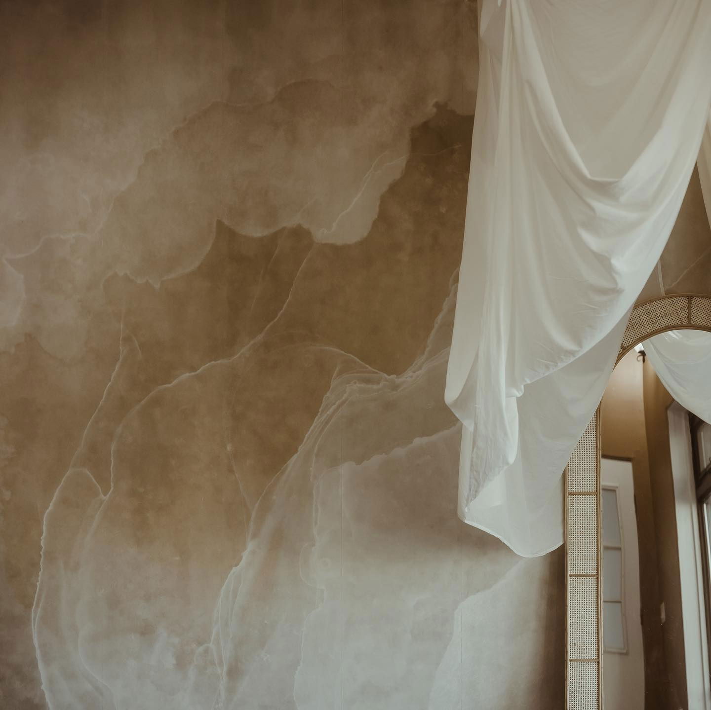 Interior con una pared con efecto de mármol marrón, cortinas blancas y un espejo de ratán.