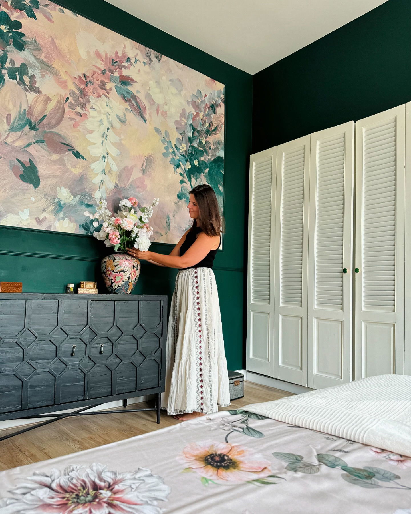 Vrouw schikt bloemen in slaapkamer met bloemenmuurkunst, grijze commode en witte luikenkast.