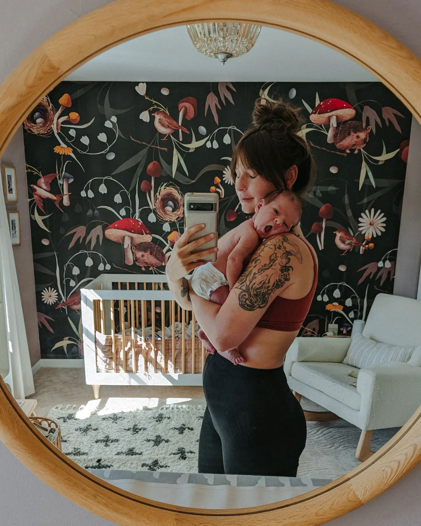 Spiegel-Selfie einer Frau, die ein gähnendes Baby in einem Kinderzimmer mit Vogel- und Pilztapete hält.