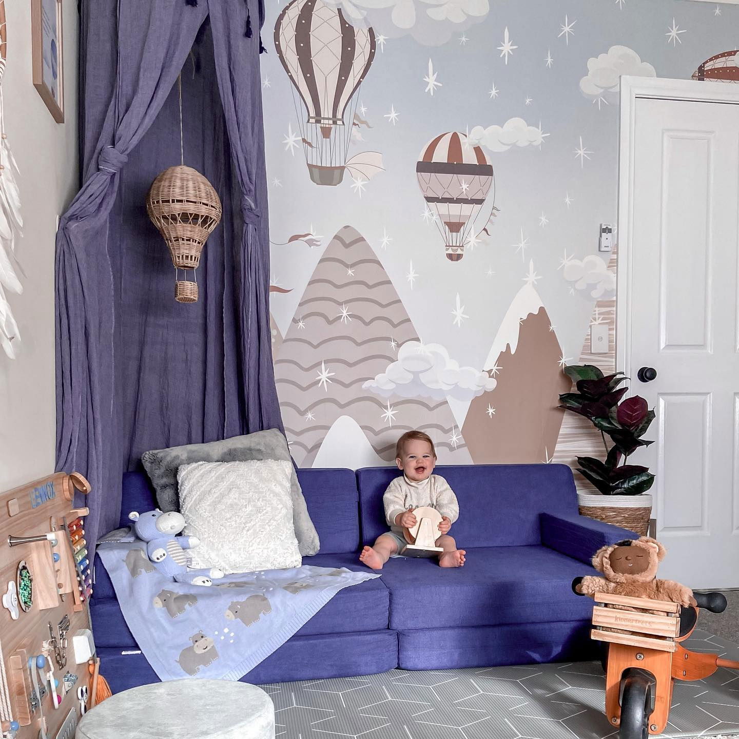 Spielzimmer-Szene mit Baby auf blauem Nugget-Sofa, das ein Holzspielzeug hält. Heißluftballon-Tapete, Baldachin und Aufsitzspielzeug.