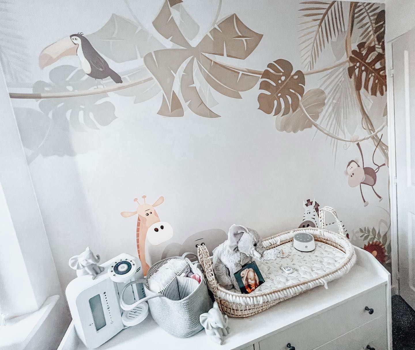 Chambre de bébé avec décoration murale sur le thème de la jungle, commode blanche et berceau en osier avec un éléphant en peluche.