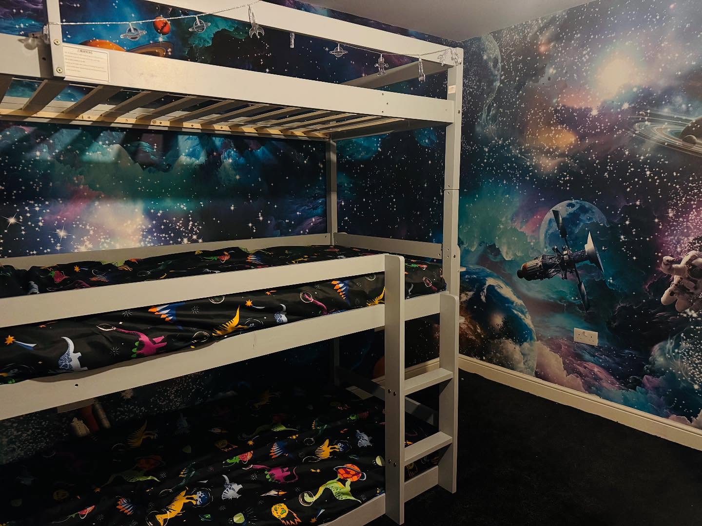 White triple bunk bed with black dinosaur print bedding, against a space-themed wallpaper.