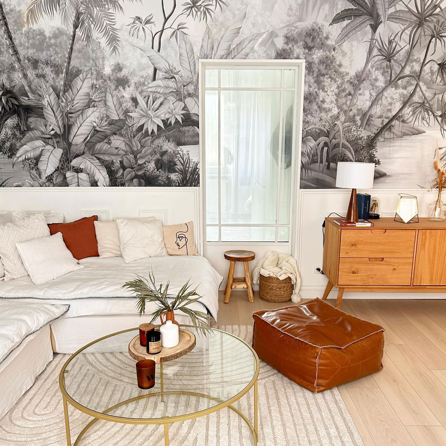 Living room with tropical mural, white sofa with pillows, glass coffee table, and brown leather ottoman.