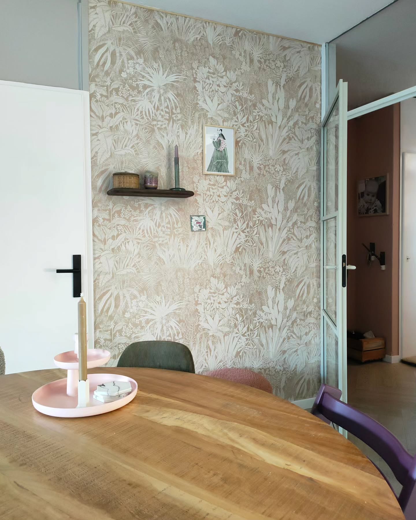 Vista interior de un comedor con una mesa de madera, un soporte para pastel rosa y papel tapiz botánico.