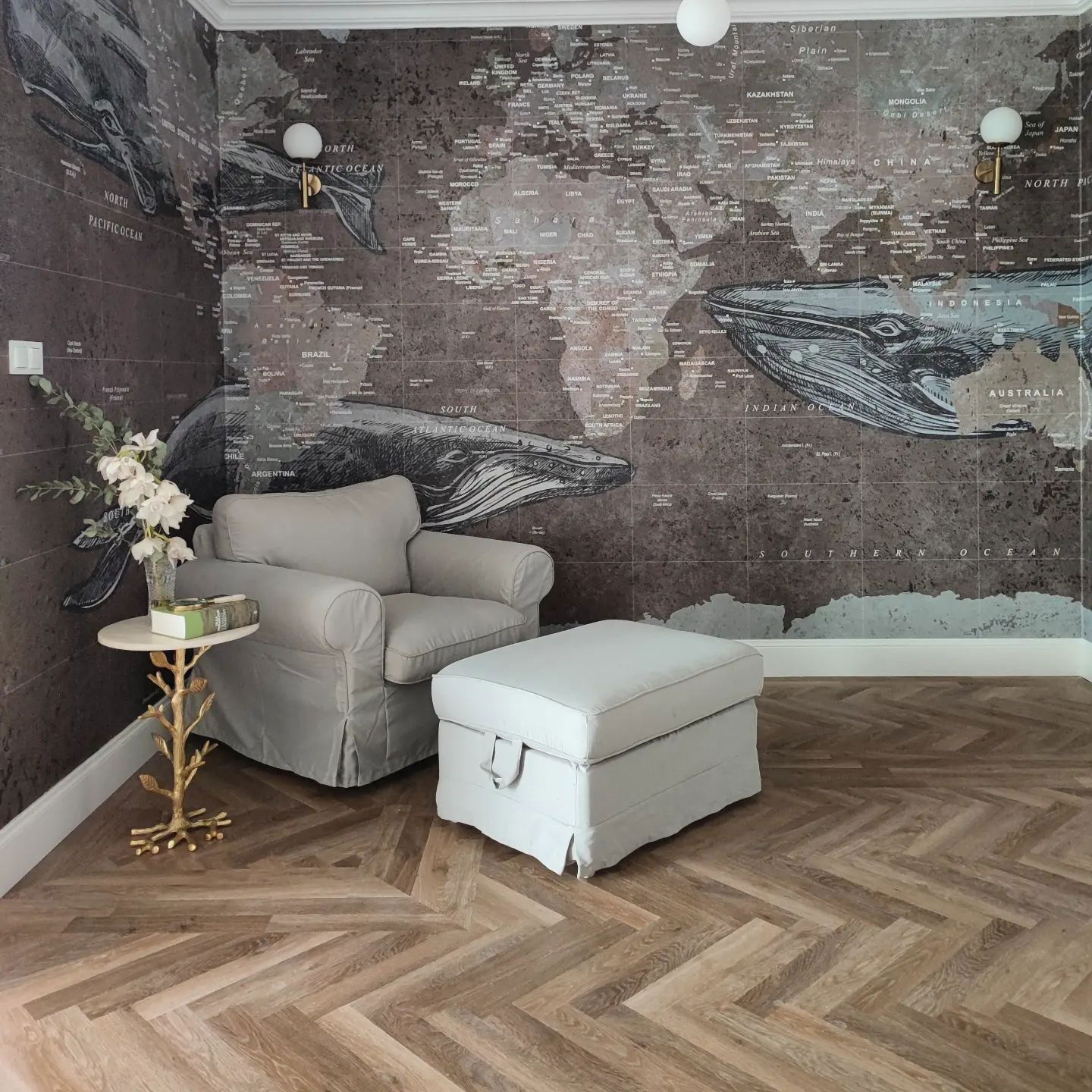 Room with world map wallpaper, gray armchair and ottoman, and herringbone wood floor.