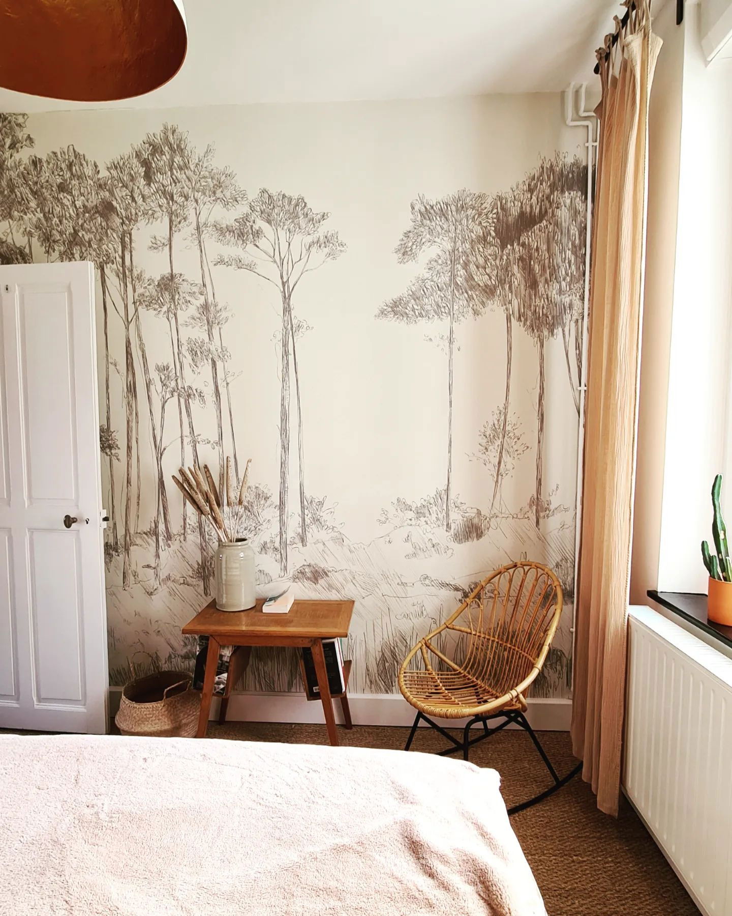 Bedroom with forest mural, rattan chair, and small wooden table with vase.
