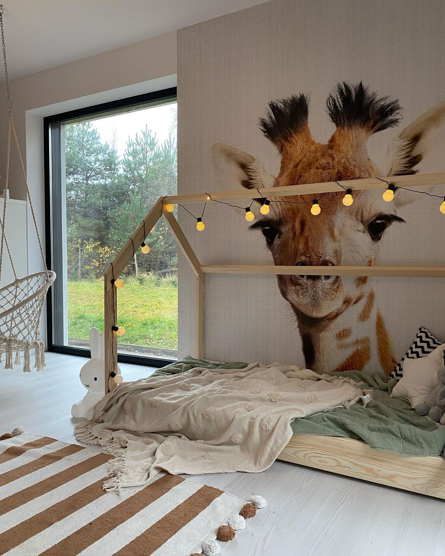 Kinderzimmer mit Hausrahmenbett, Giraffen-Wandbild und gestreiftem Teppich.