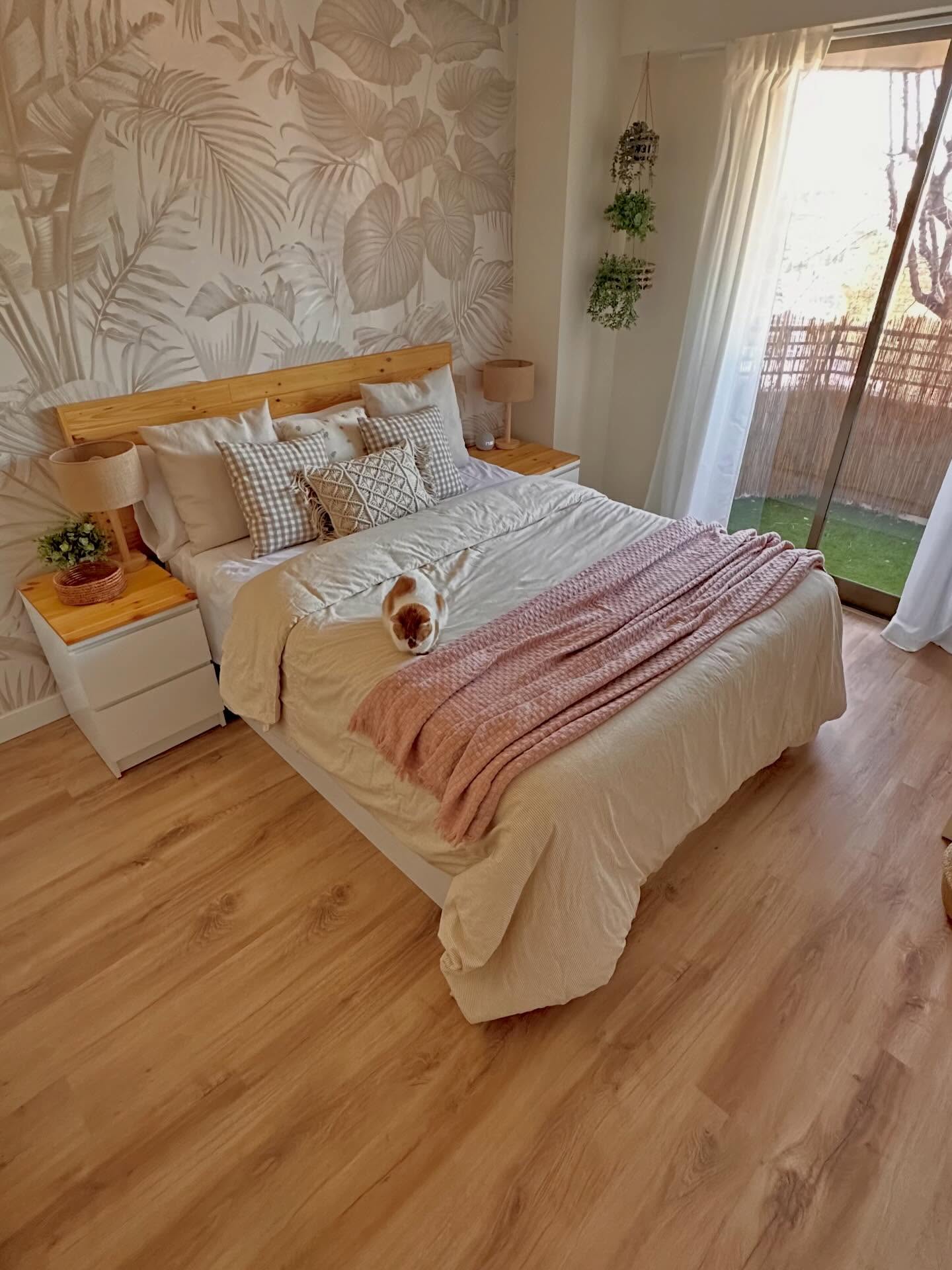 A light-filled bedroom with a cat resting on a cream bed with a pink throw. It has a light wood headboard, two nightstands with lamps, leaf-patterned wallpaper, and light wood floors.