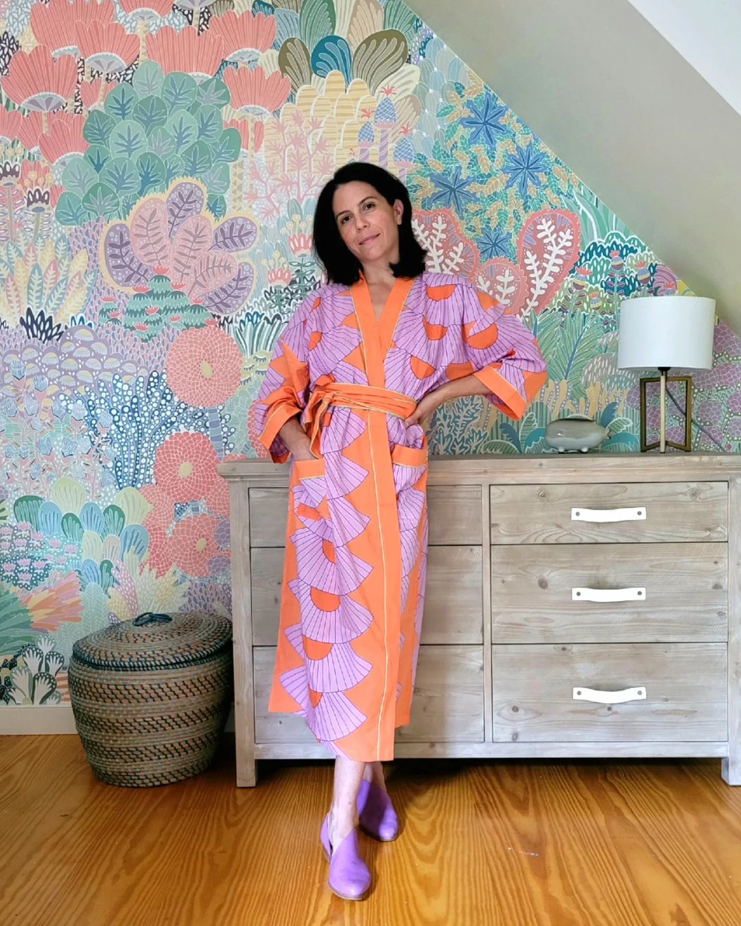 Woman in orange robe with purple accents, standing in front of a colorful floral wallpaper and wooden dresser.