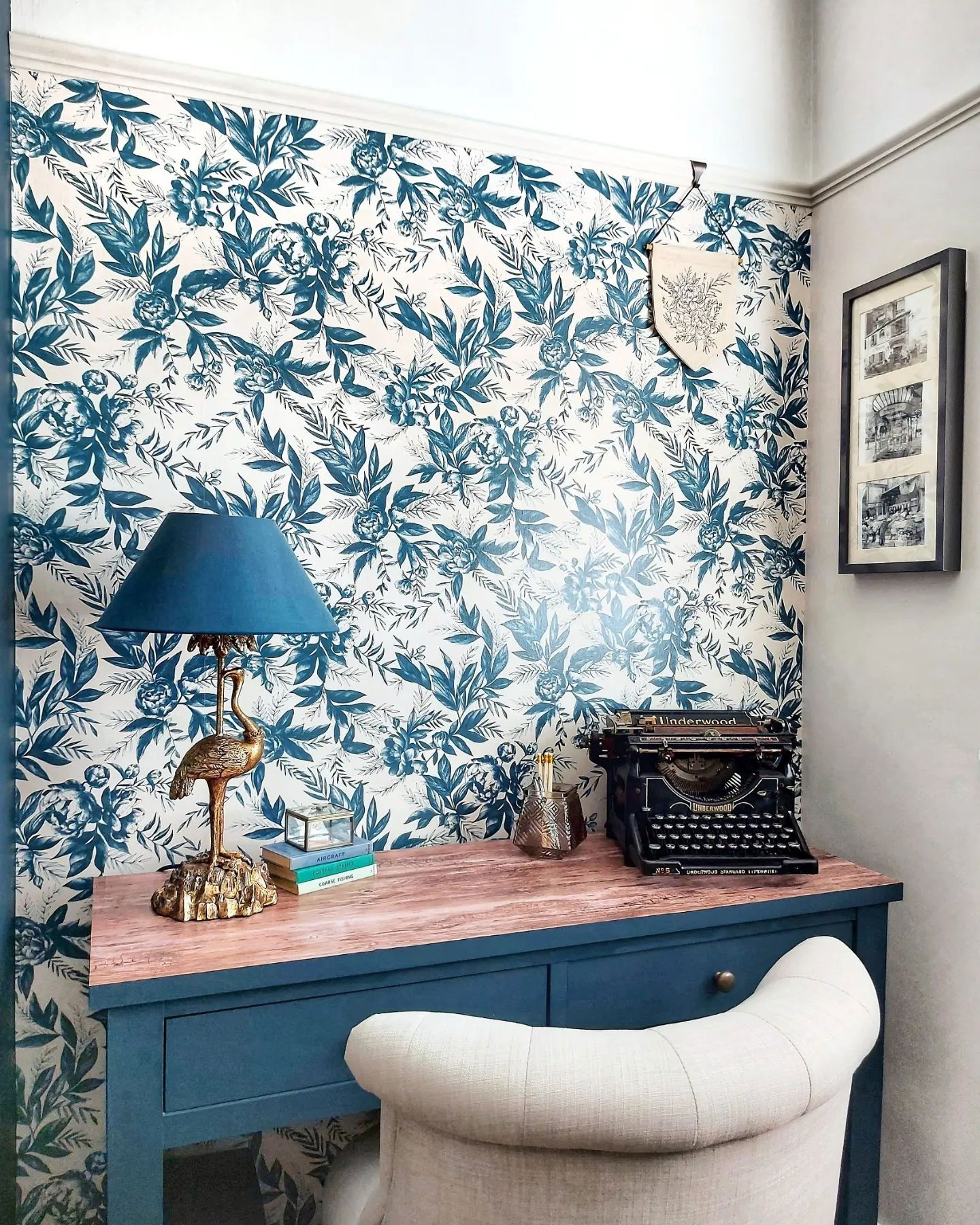 Vintage desk with Underwood typewriter, gold lamp, and floral wallpaper.