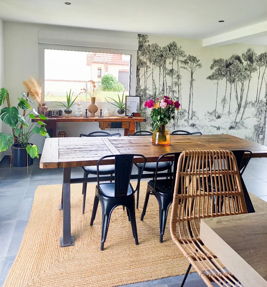 Esszimmer mit Holztisch, schwarzen Metallstühlen und gewebtem Stuhl auf Jute-Teppich. Waldgemälde an der Wand.