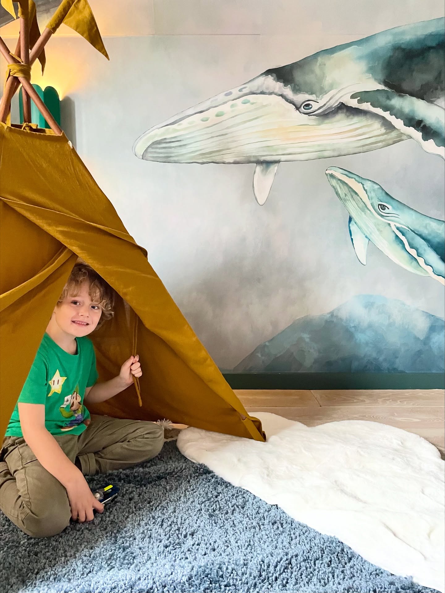 A young child smiles from inside a mustard-colored teepee in a room with a whale mural.