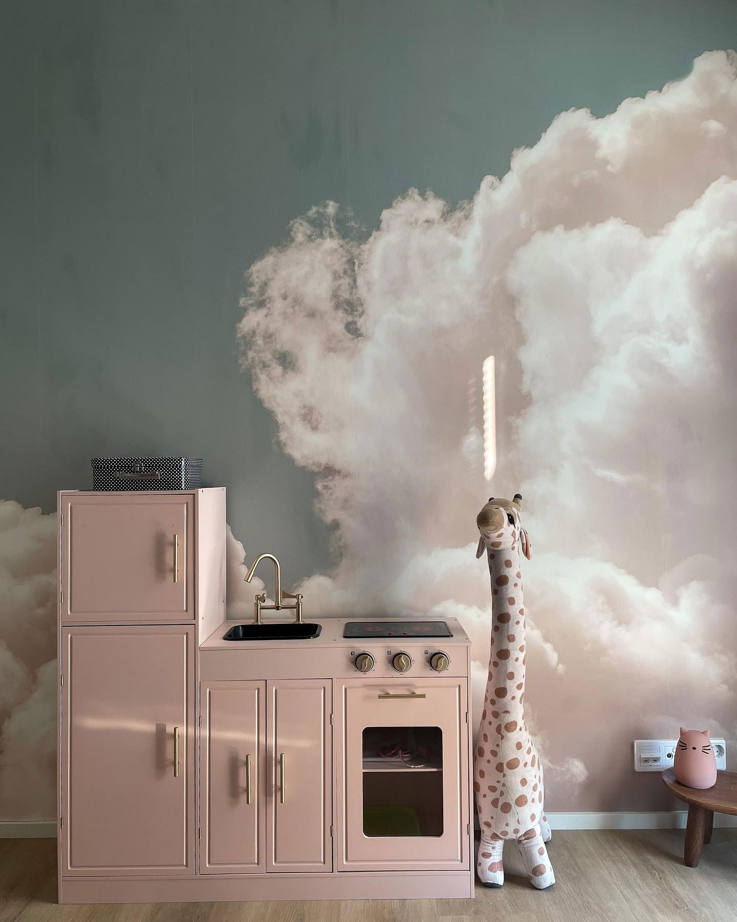 Playroom with pink toy kitchen, stuffed giraffe, and cloud wallpaper.