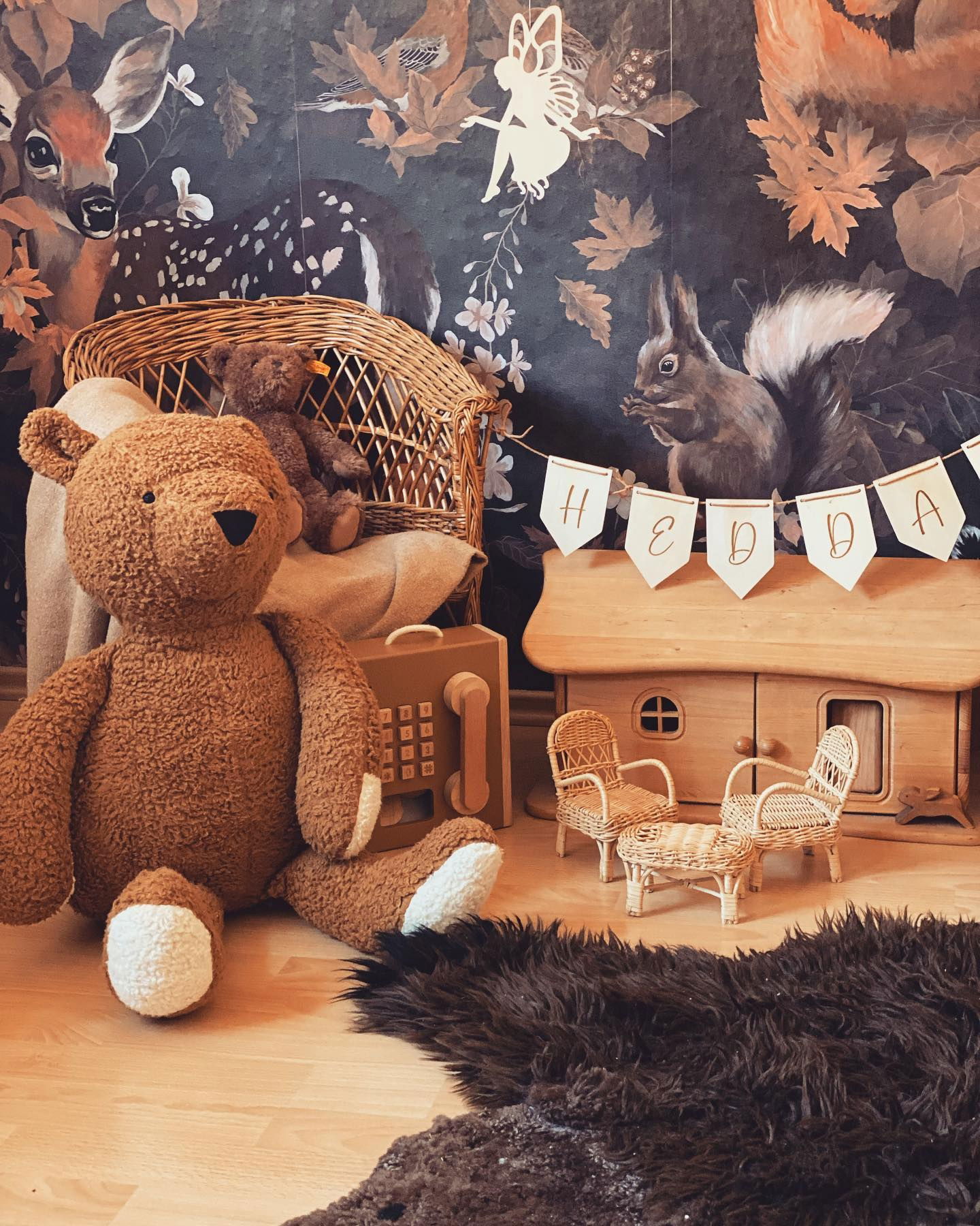 Playroom scene with large teddy bear, dollhouse, and woodland wallpaper.