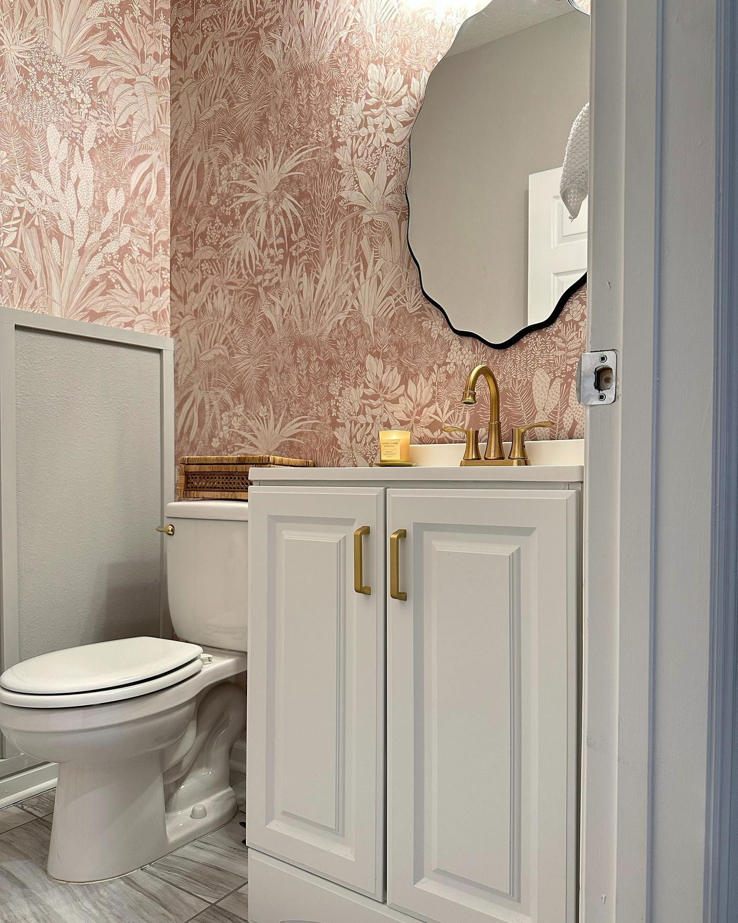Baño con papel tapiz floral rosa, tocador blanco con grifo dorado y espejo irregular con marco negro.
