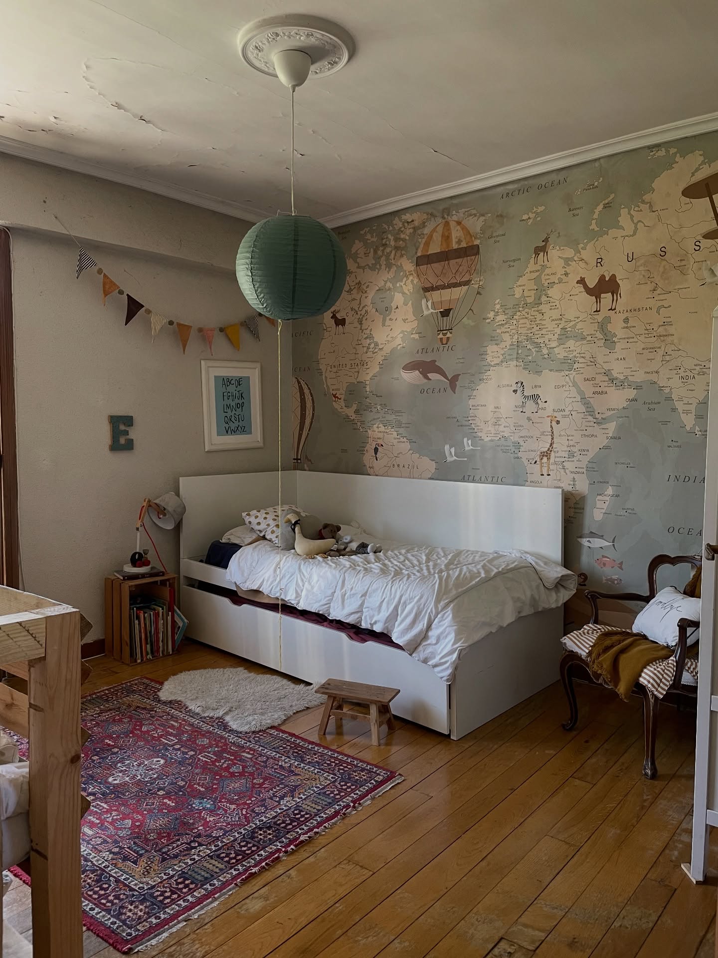 Camera da letto per bambini con mappa del mondo sul muro, letto bianco, tappeto a motivi rossi e lanterna di carta verde.