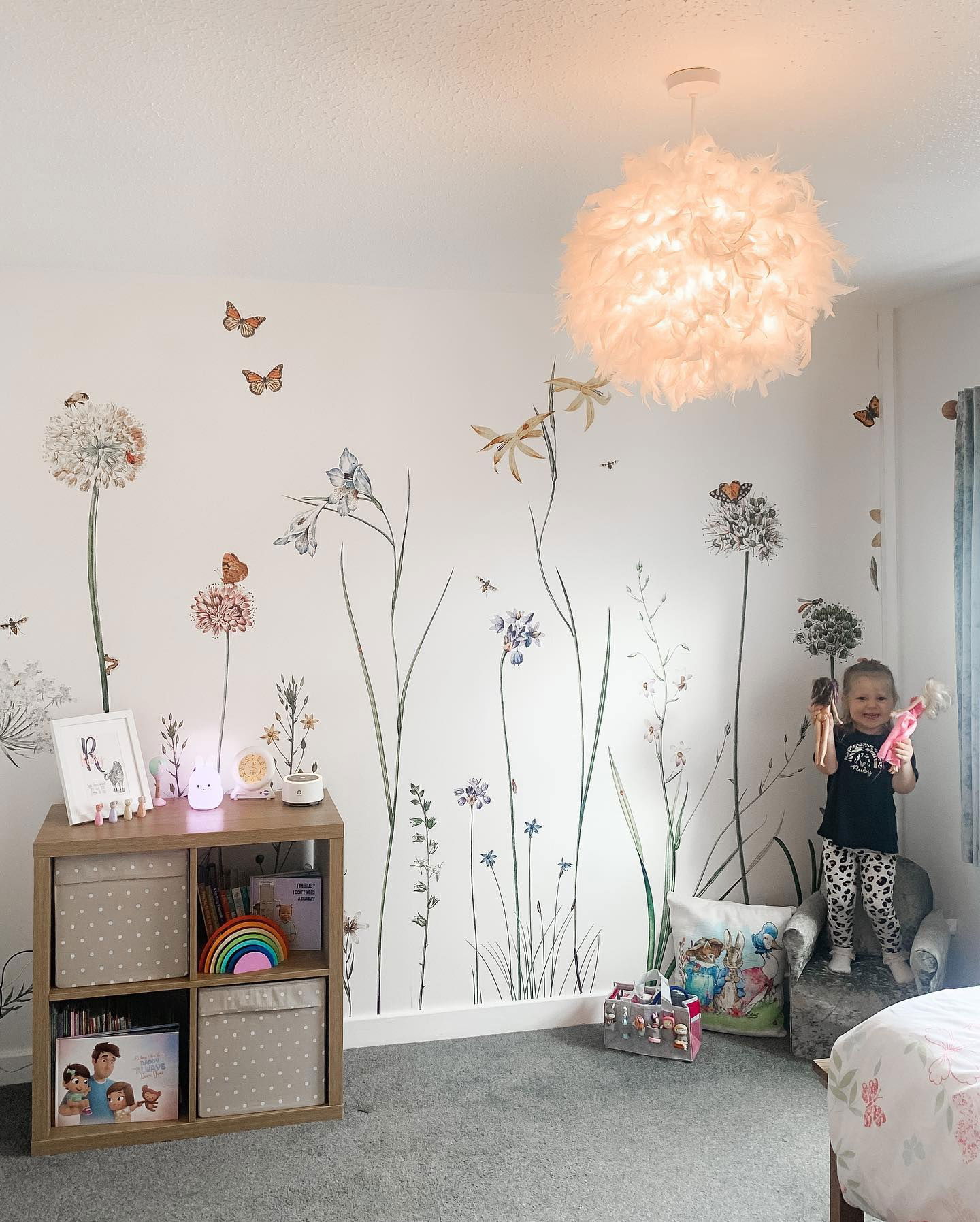 Enfant debout sur une chaise dans une chambre à thème floral, tenant des poupées. La chambre comprend des autocollants muraux de fleurs, un luminaire en plumes et une unité de rangement cubique.