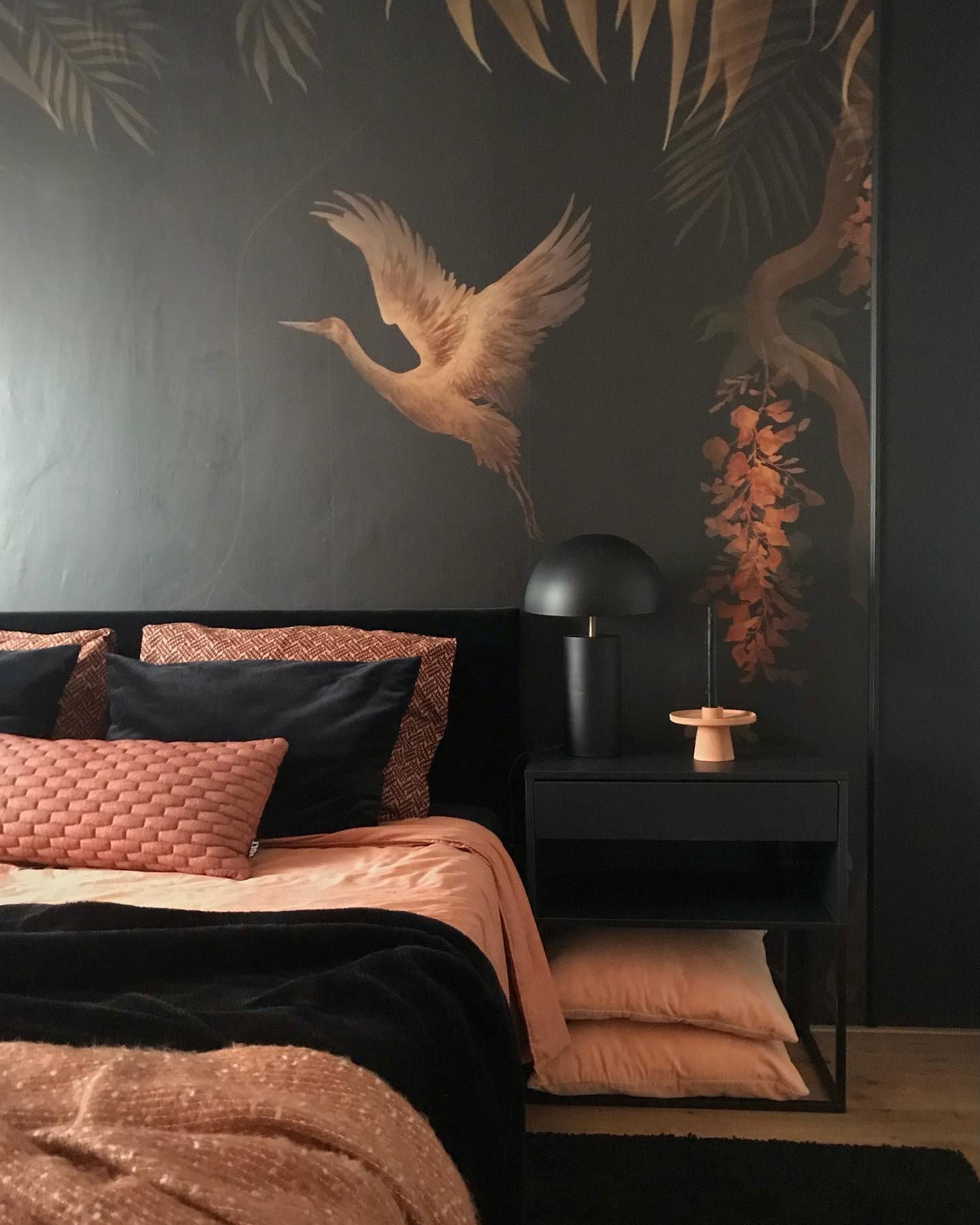 Schlafzimmer mit schwarzem Kopfteil, pfirsichfarbener Bettwäsche und Nachttisch mit Lampe. Die Wandkunst zeigt einen Kranich und Blätter.