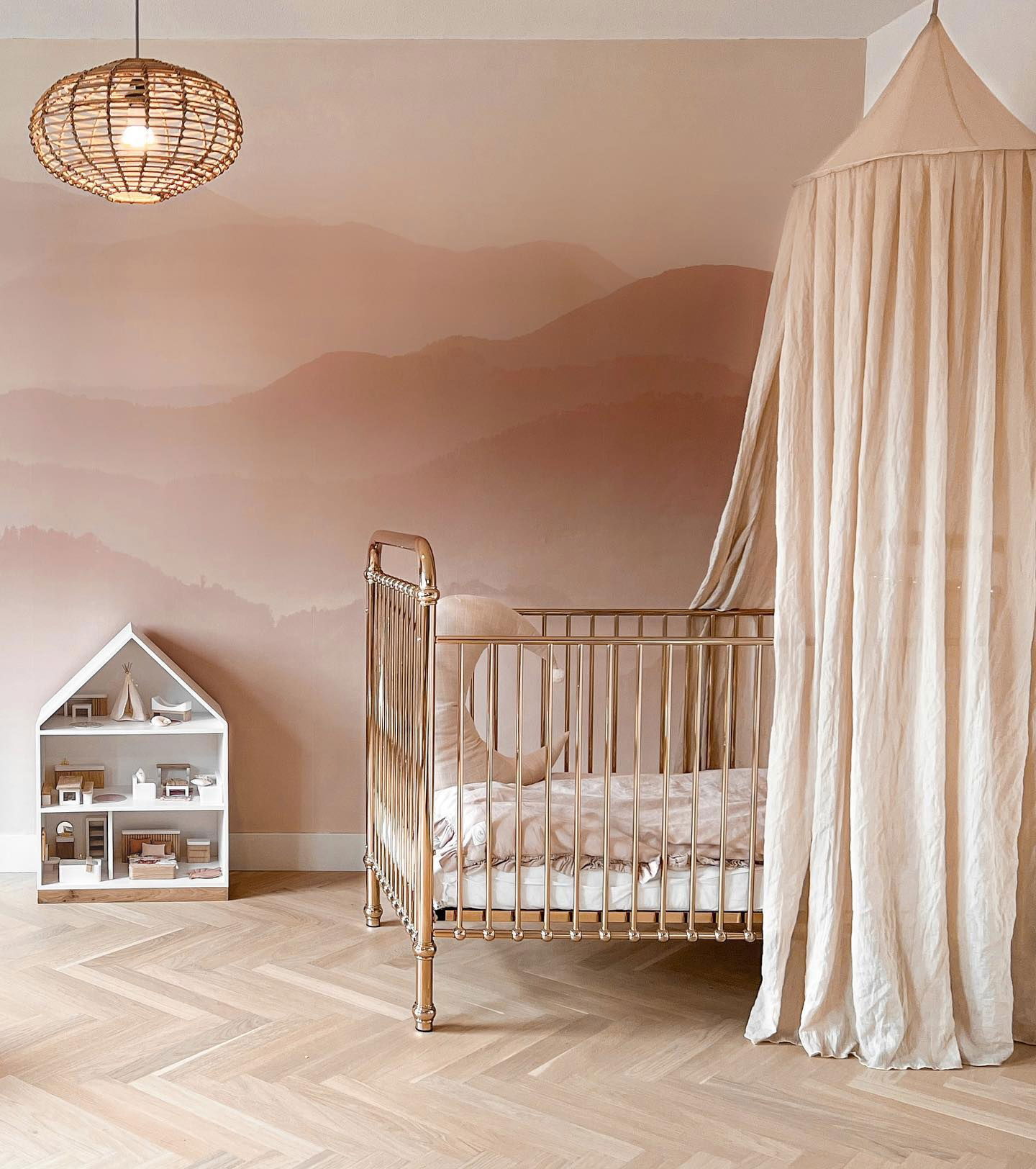 Nursery with gold crib, canopy, and mountain mural. Rattan pendant light and white dollhouse.