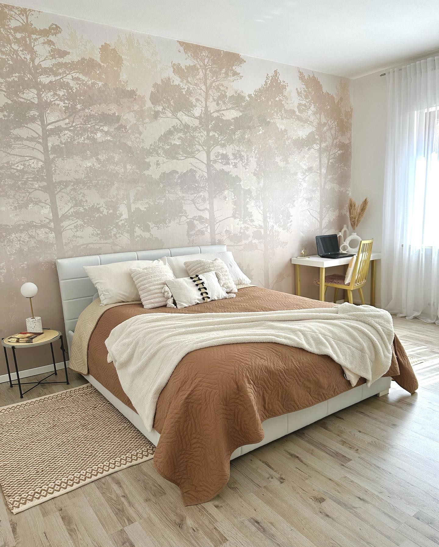 Bedroom with white bed, brown quilt, and forest wallpaper. Desk with laptop and gold chair.