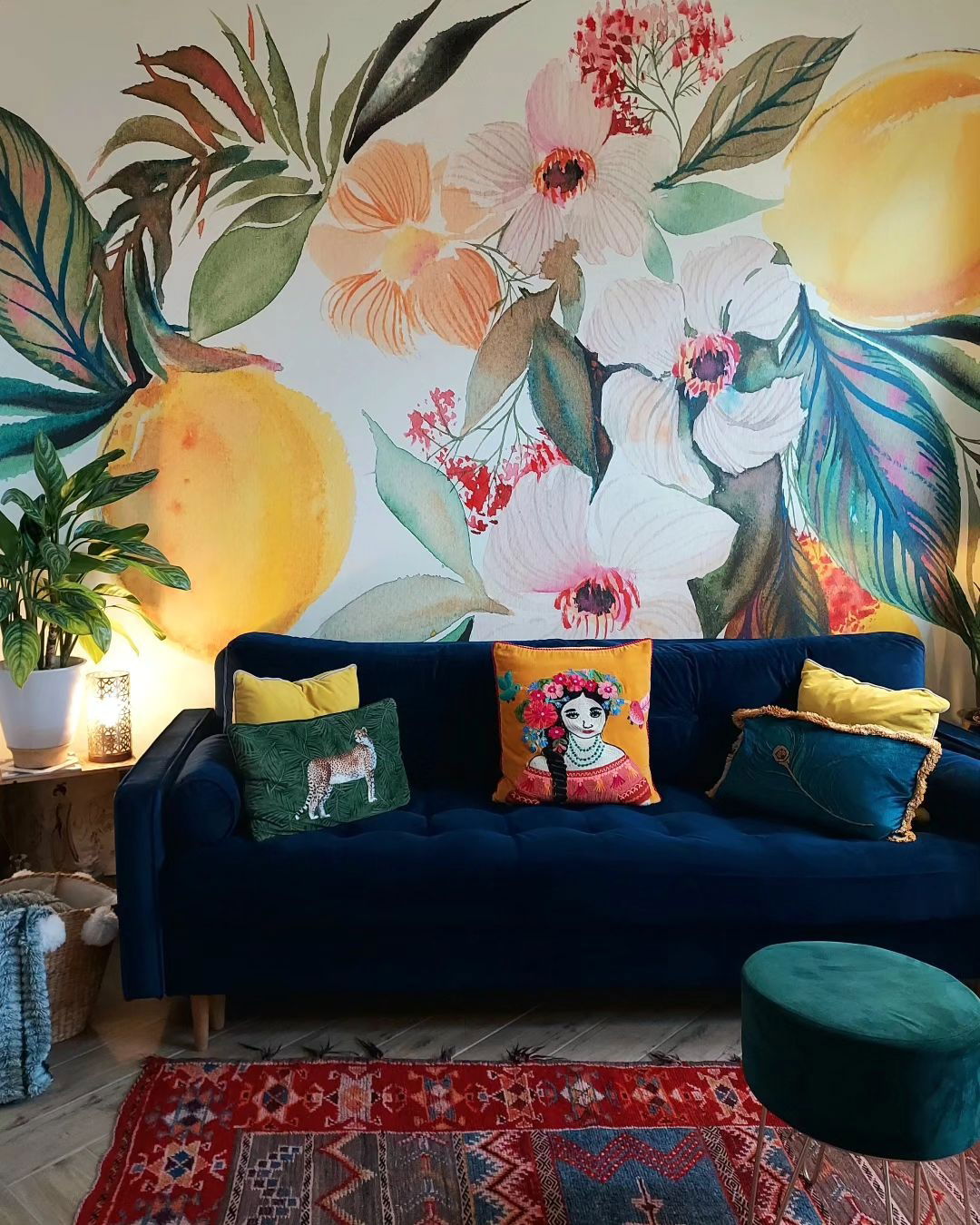 Living room with blue velvet sofa, colorful pillows, floral mural, and patterned rug.