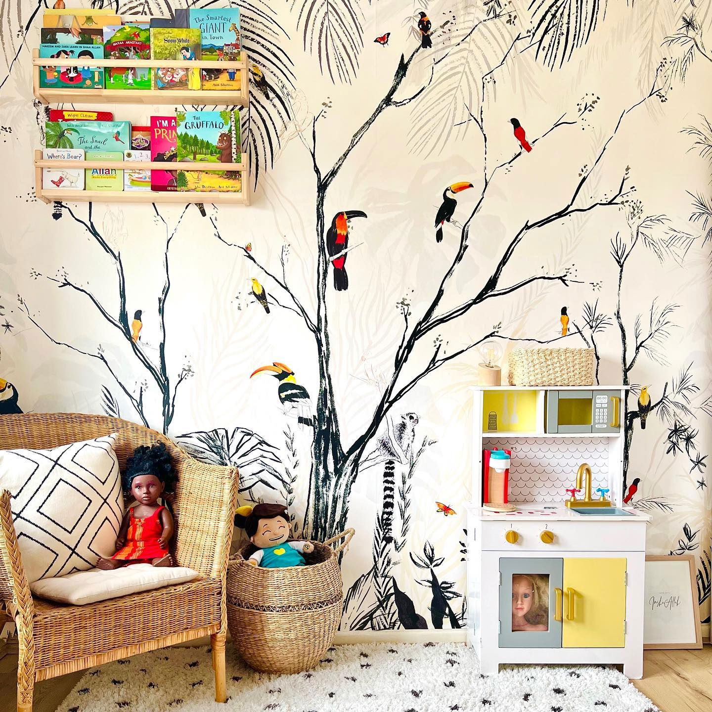 Child's room with jungle wallpaper, wooden toy kitchen, and wicker chair with dolls. Bookshelf with children's books on the wall.