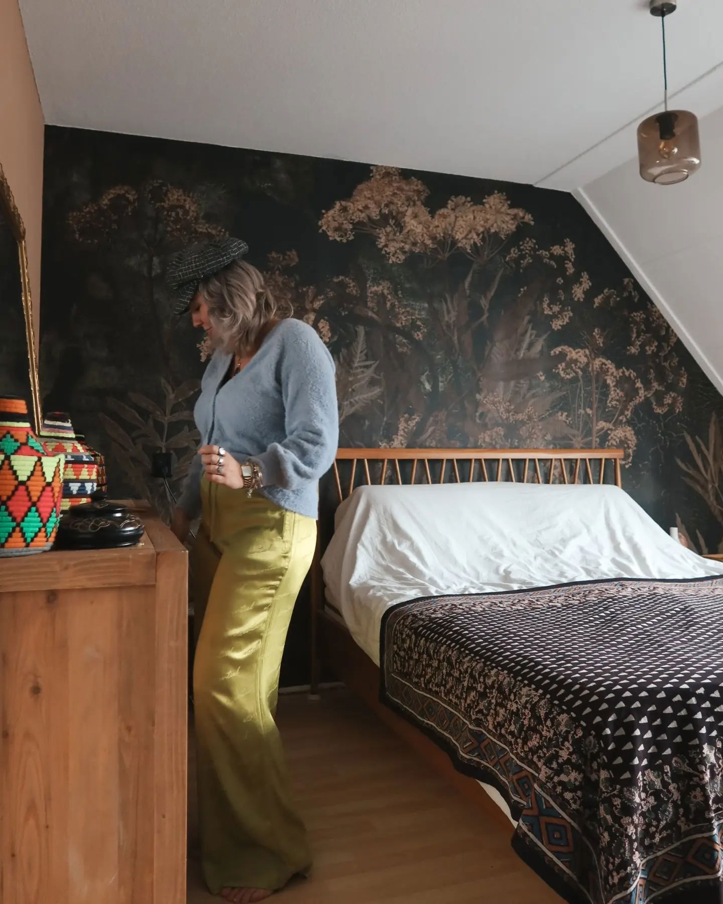 Eine Frau in einer blauen Strickjacke und chartreusefarbenen Hose steht in einem Schlafzimmer mit floraler Tapete.