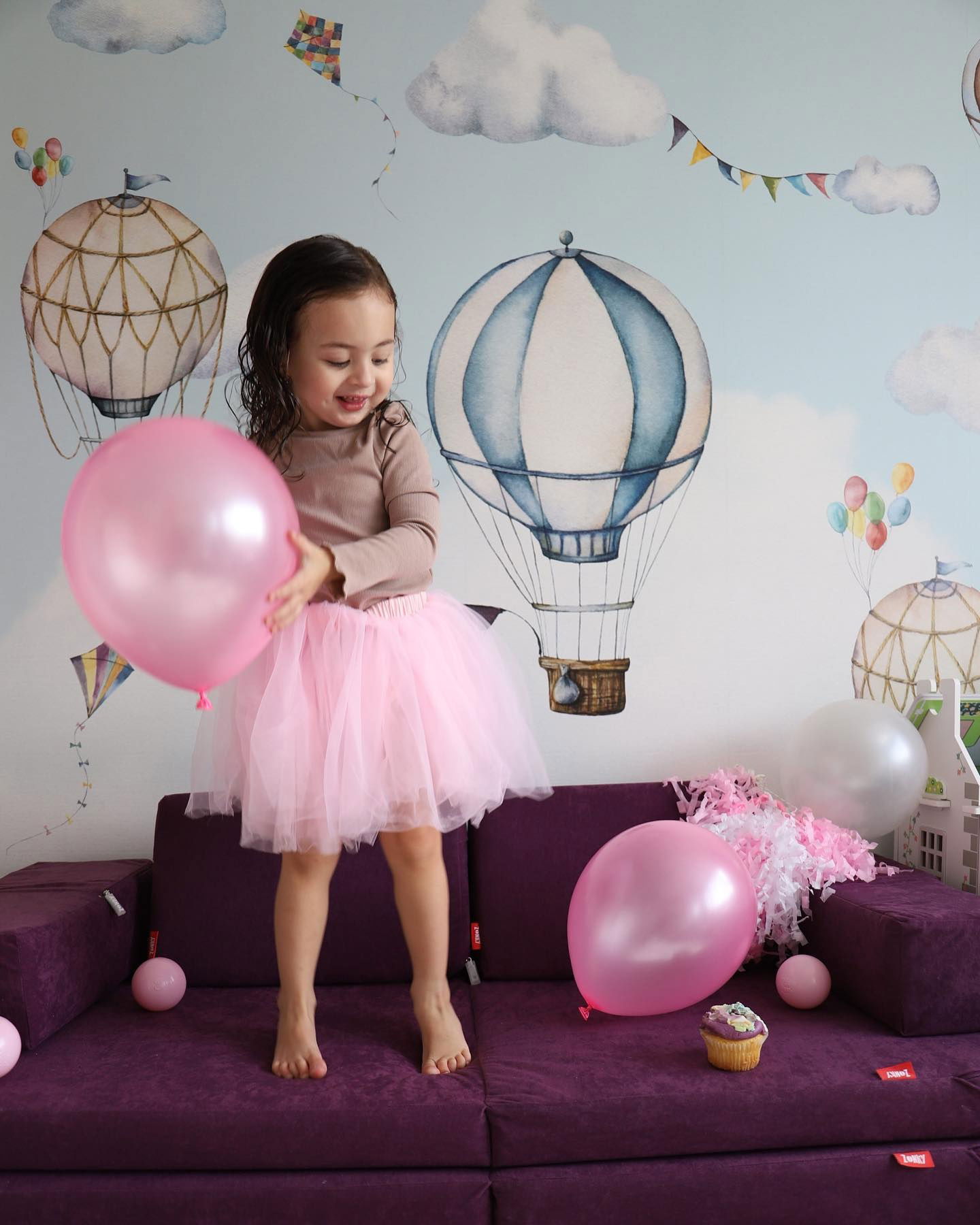 A young girl in a pink tutu plays with balloons on a purple Nugget couch.